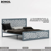 A promotional image of the "ARISTORA Metal Bed" by Bongo, presented in a grey and white color scheme. The modern bed frame is made of slate-grey metal. The tall, rectangular headboard and matching lower footboard feature a decorative laser-cut pattern of organic, pebble-like shapes. The grey cutout panel is backed by a solid white panel, creating a high-contrast, two-tone effect. The bed is made with dark brown and black bedding and is set against a plain, light grey background.